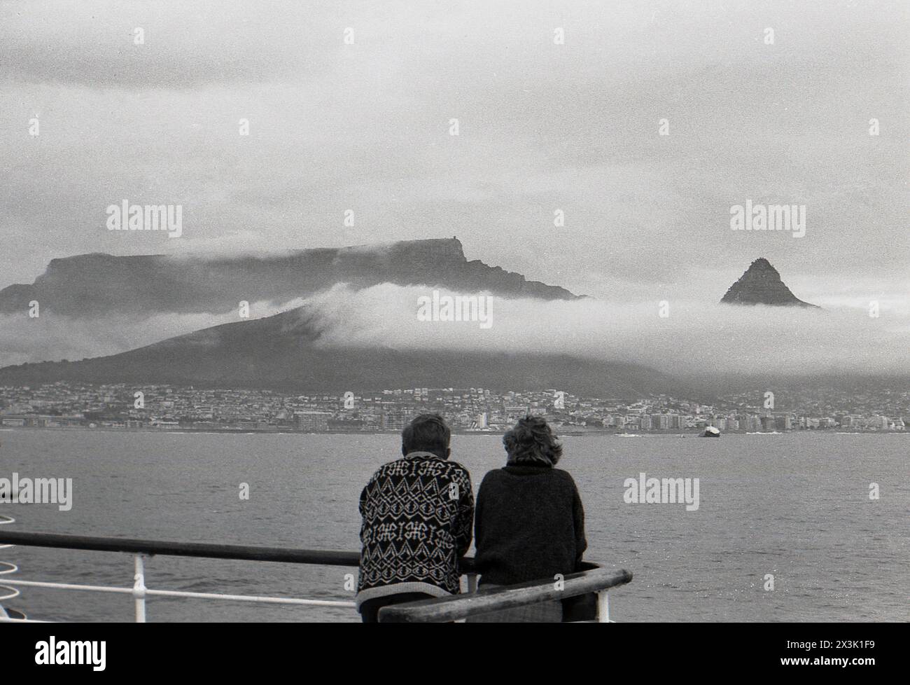 1960er Jahre, historisch, zwei Passagiere stehen auf dem Balkon eines Kreuzfahrtschiffes mit Blick auf die Skyline des Tafelbergs, auf der Kap-Halbinsel Südafrika, Wolken über den Bergen. Stockfoto