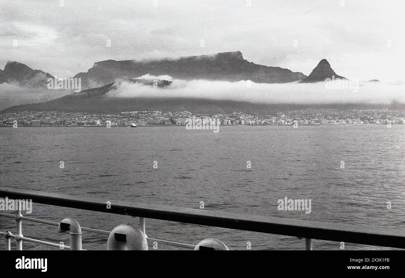 1960er Jahre, historisch, zwei Passagiere stehen auf dem Balkon eines Kreuzfahrtschiffes mit Blick auf die Skyline des Tafelbergs, auf der Kap-Halbinsel Südafrika, Wolken über den Bergen. Stockfoto