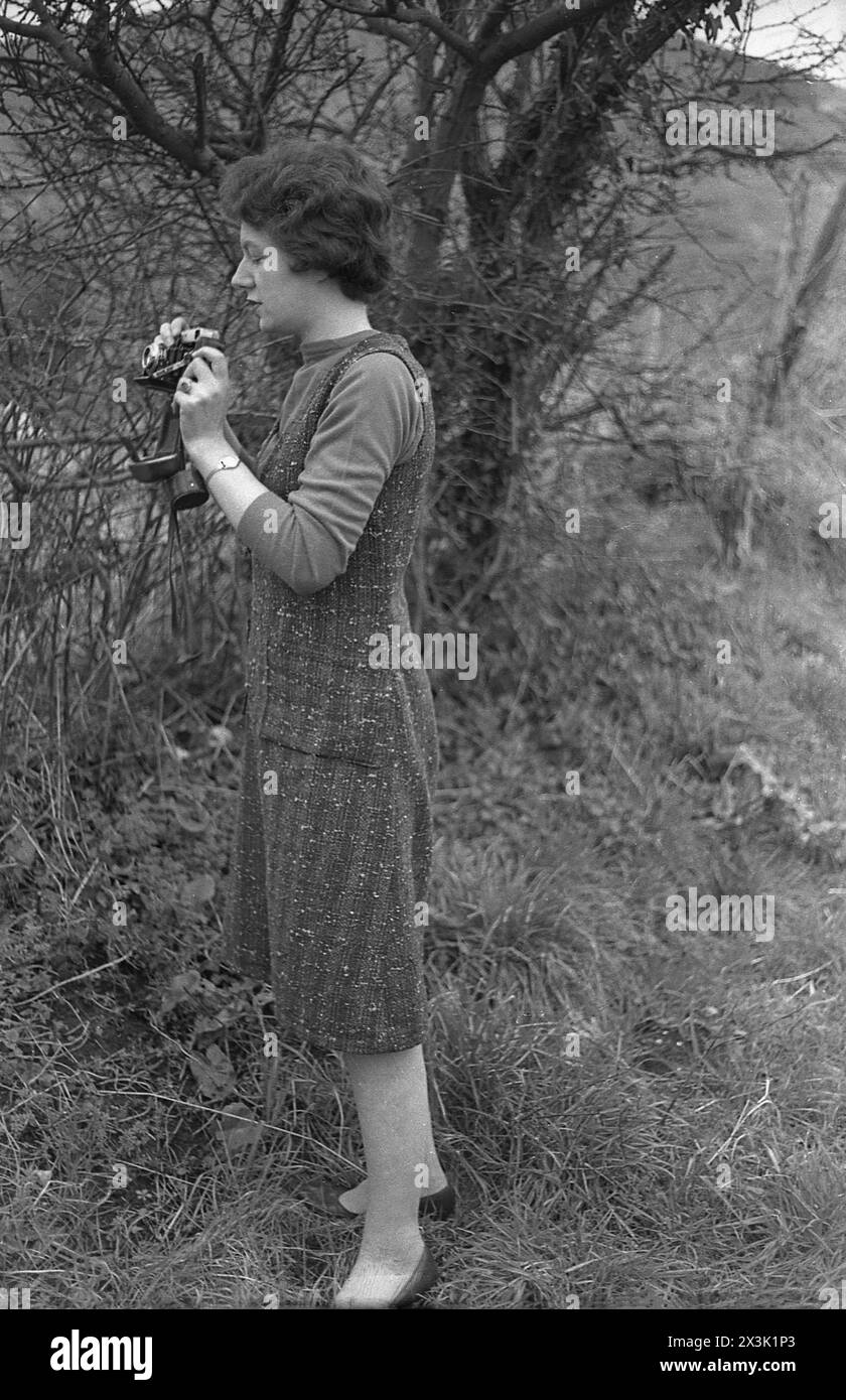 1960er Jahre, historisch, draußen an einem ländlichen Grasrand, eine junge Dame in einem halbwadenlangen Kleid, stehend mit einer faltbaren Filmkamera aus dieser Zeit. Stockfoto