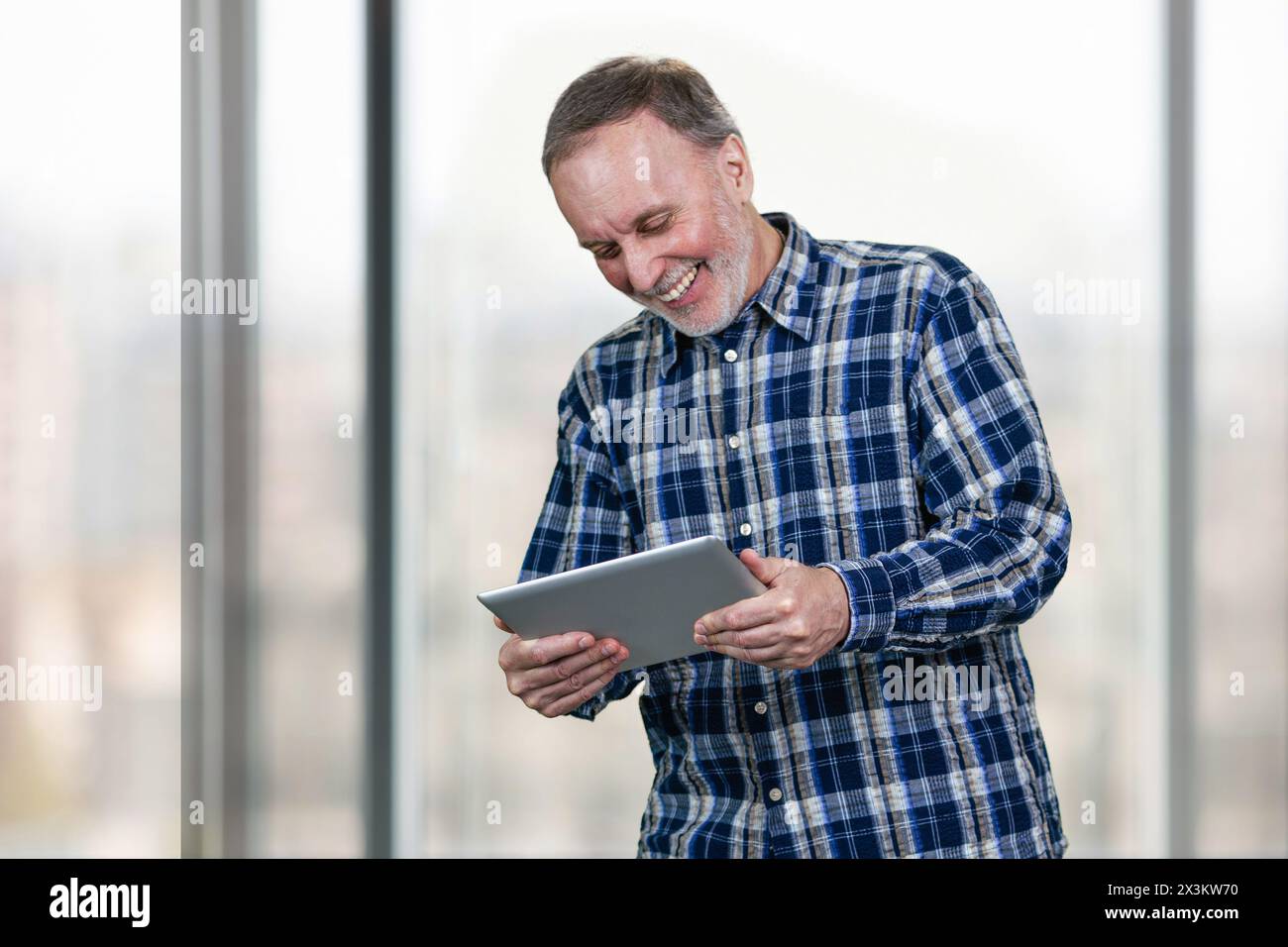 Lächelnder reifer Mann, der den Tablet-pc in beiden Händen hält. Riesige Bürofenster im Hintergrund. Stockfoto