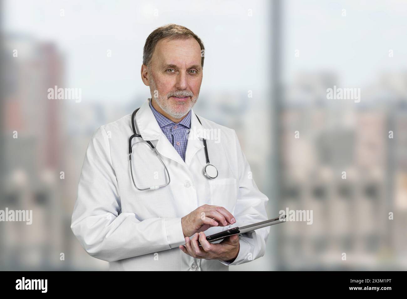 Porträt eines reiferen männlichen Arztes, der ein digitales Tablet hält, das drinnen steht. Abstrakter Hintergrund für verschwommene Fenster. Stockfoto