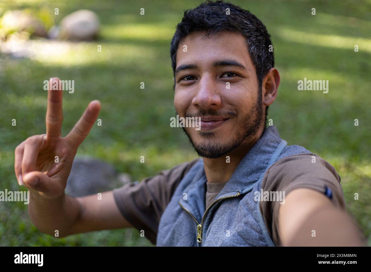 Selbstporträt eines jungen lateinamerikanischen Erwachsenen (24) macht eine positive winkende Geste vor seinem Handy. Technologiekonzept. Stockfoto