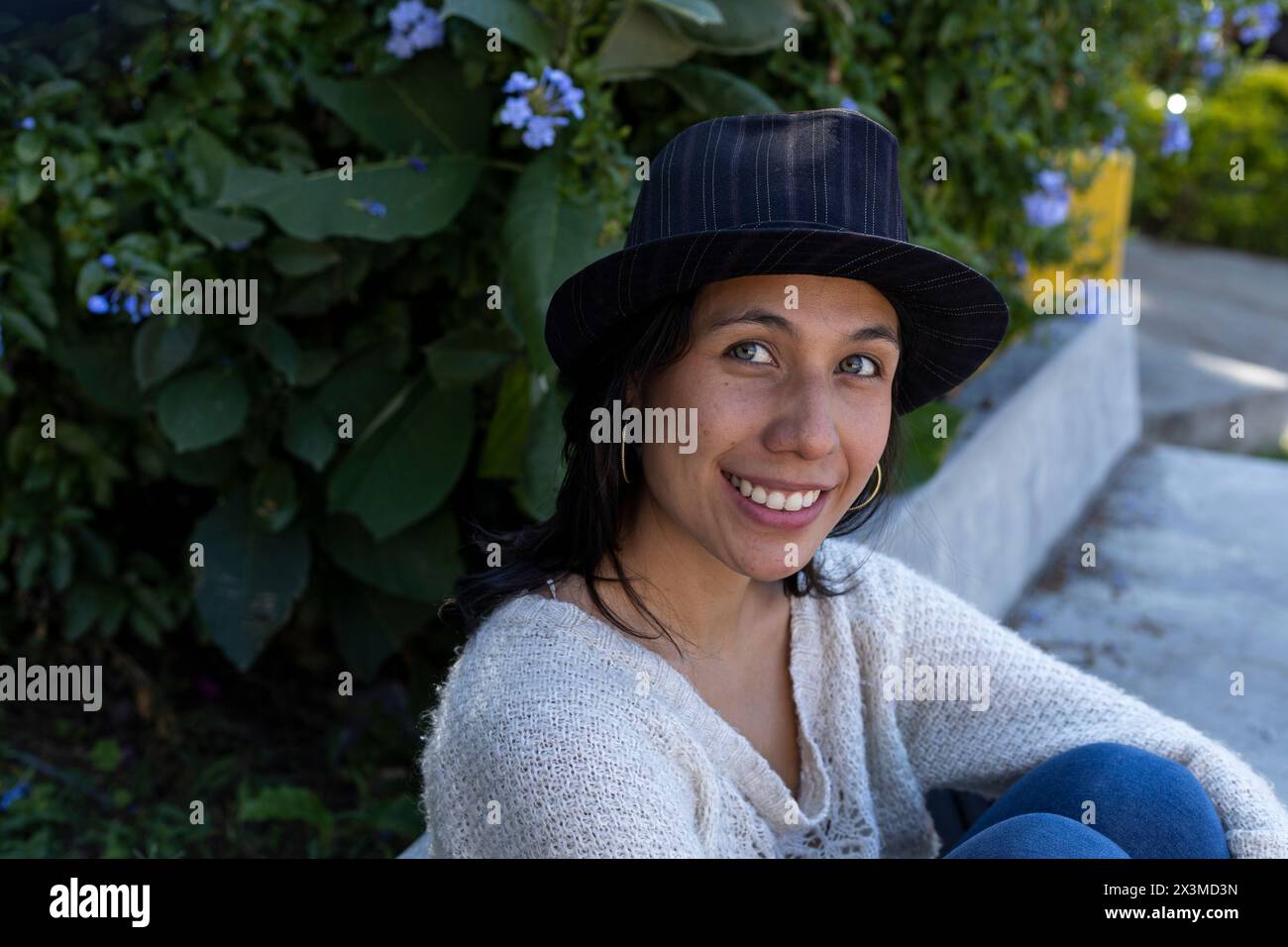 Lateinamerikanische Erwachsene Frau (29) mit grünen Augen, sie sitzt im Park und schaut mit einem Lächeln und positiver Einstellung in die Kamera, trägt einen Hut. L Stockfoto