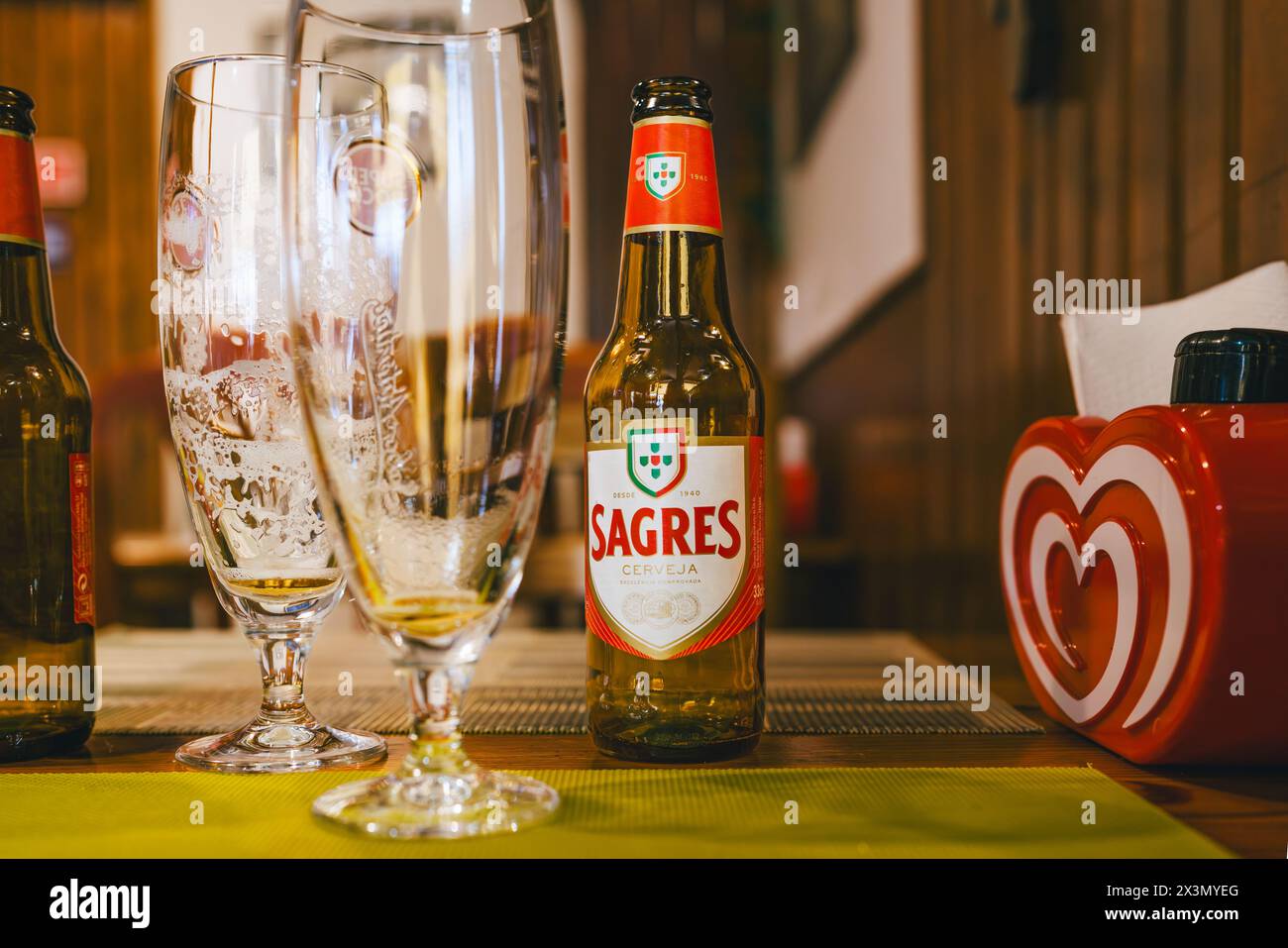 Tavira, Portugal, 8. Januar 2024. Sagres Bierflasche auf einem Tisch mit leeren Biergläsern und einem roten Serviettenspender in Herzform in gemütlicher Umgebung Stockfoto
