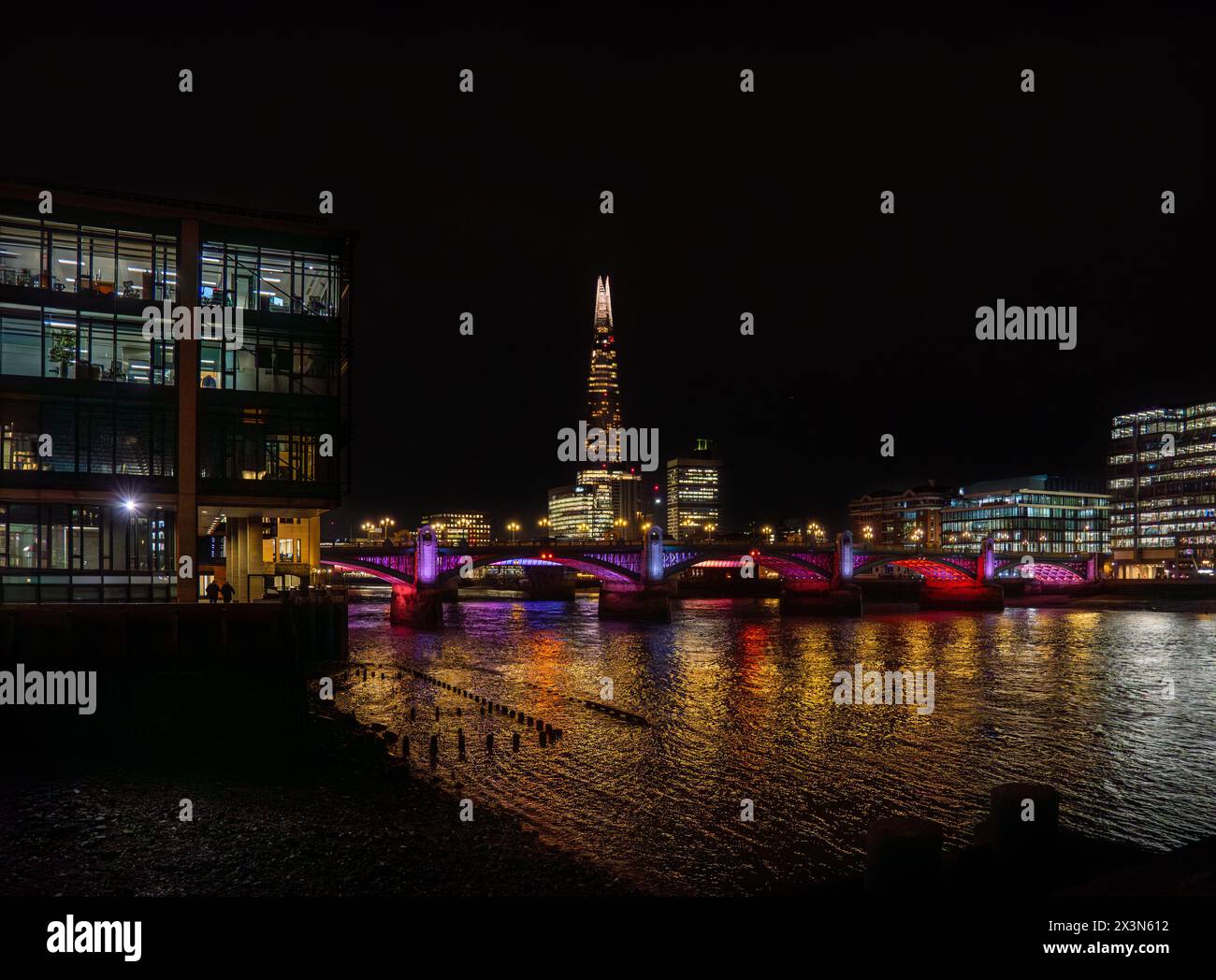 Die Blackfriars Bridge ist nachts mit violetten Lichtern beleuchtet, die ihre Lichter auf der Themse und ihrem Ufer reflektieren, mit dem Shard-Wolkenkratzer hinter einem Stockfoto