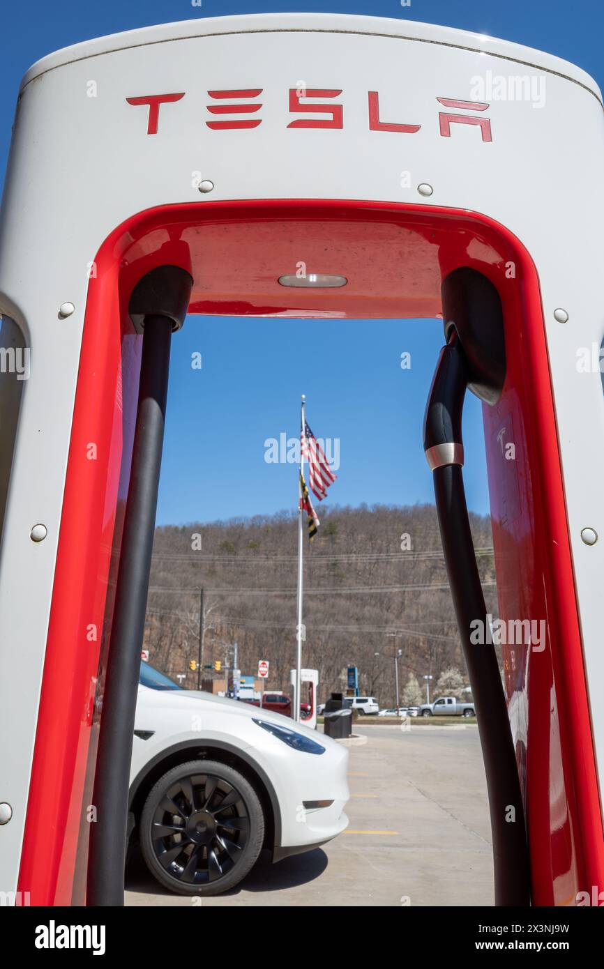 La Vale Maryland USA-03 29 2024: In La Vale, Maryland, USA, stürmt ein weißes Auto an einem Tesla Supercharger unter der flatternden amerikanischen Flagge gegen A Stockfoto