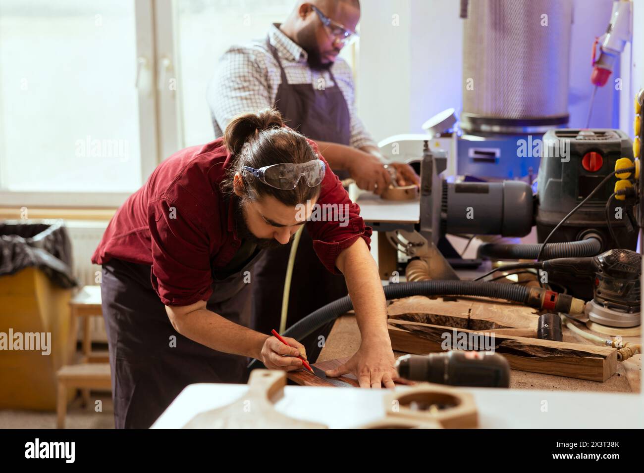 Holzarbeiter mit Lineal zur Messung von Holzdielen mit einem afroamerikanischen Kollegen. Kreative Person, die mit Bleistift auf Holzbrett zeichnet, um zu bestimmen, wo der Schnitt gemacht werden soll, unterstützt durch den Auszubildenden Stockfoto
