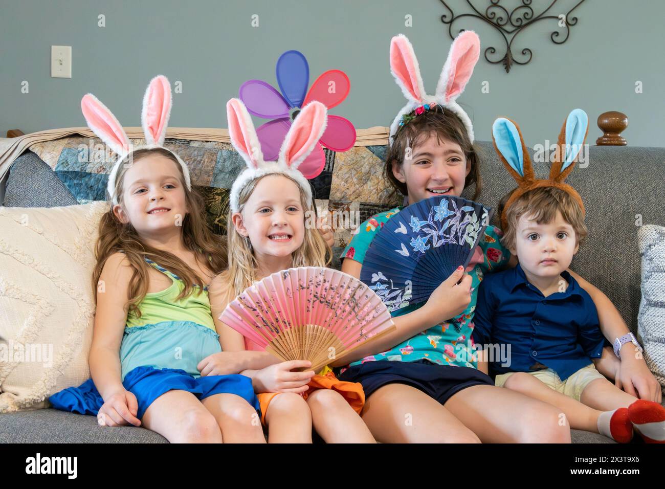 Eine Familie mit drei Mädchen, Schwestern und einem Kleinkind, Bruder sitzt auf einer Couch, zeigt ihre osterhasenohren auf dem Kopf und lächelt. Stockfoto
