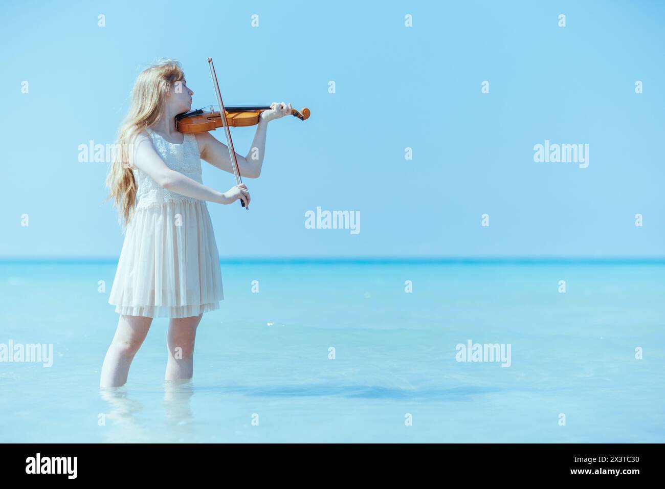 Modernes Teenager-Mädchen in weißem Kleid mit Violine, das am Meer spielt. Stockfoto