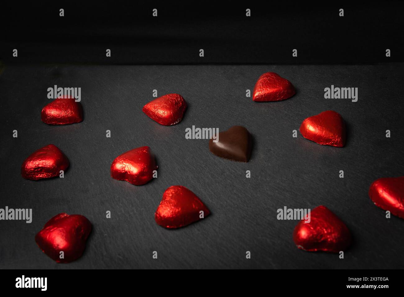 Gruppe herzförmiger Pralinen mit roter Verpackung, eine davon ohne Verpackung auf schwarzem Hintergrund mit Kopierraum. Chocolate Love Konzept Stockfoto