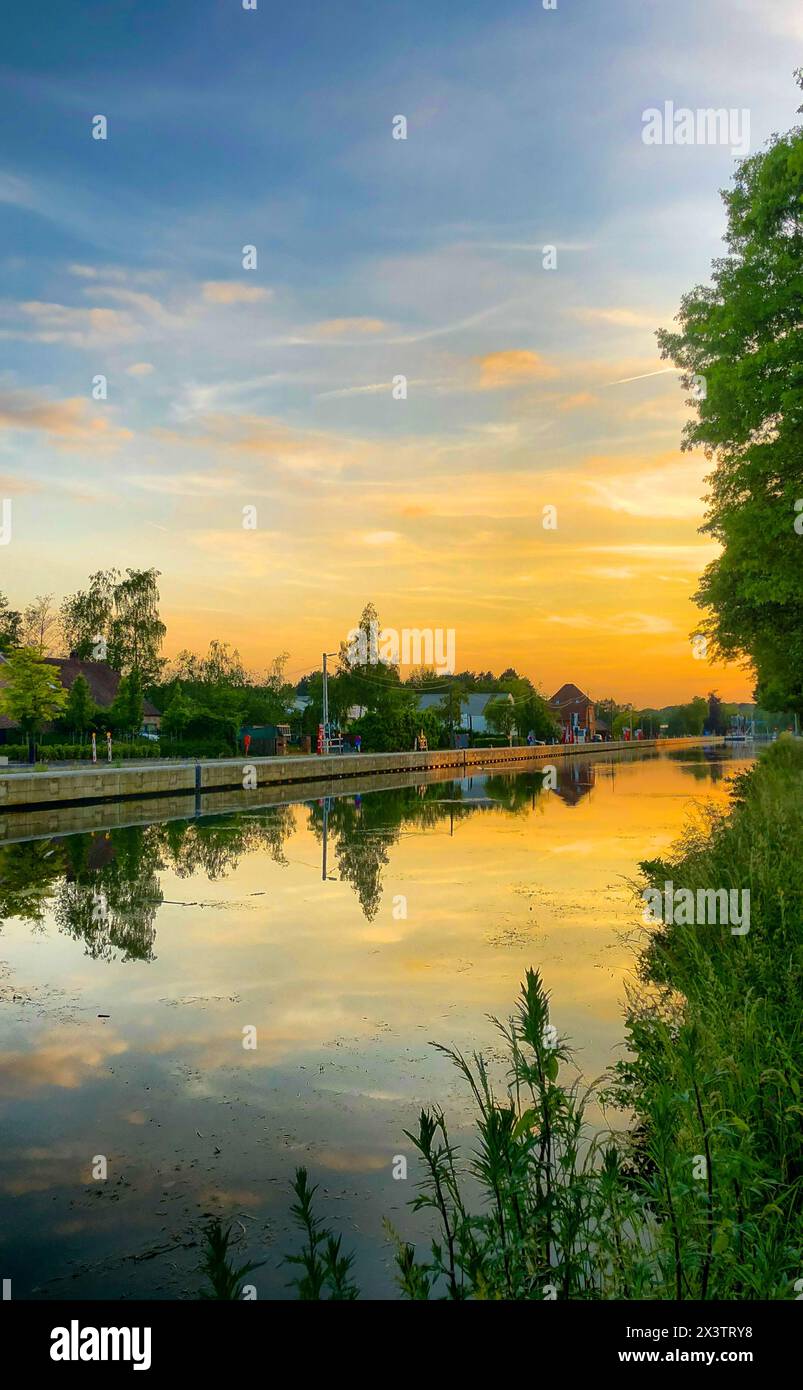 Dieses Bild zeigt eine ruhige Kanalszene bei Sonnenuntergang. Die Wasserstraße spiegelt den Himmel mit Orangen- und Blautönen und einem verlaufenden Himmel wider. Flora umrahmt den Rand des Wassers. Im Hintergrund ist eine kleine Dorfsilhouette mit Bäumen und Gebäuden unter dem weichen Himmel zu sehen, die eine ruhige Abendatmosphäre erzeugt. Ruhiger Sonnenuntergang am Kanal: Reflektierender Wasserweg mit Flora und Village Silhouette. Hochwertige Fotos Stockfoto