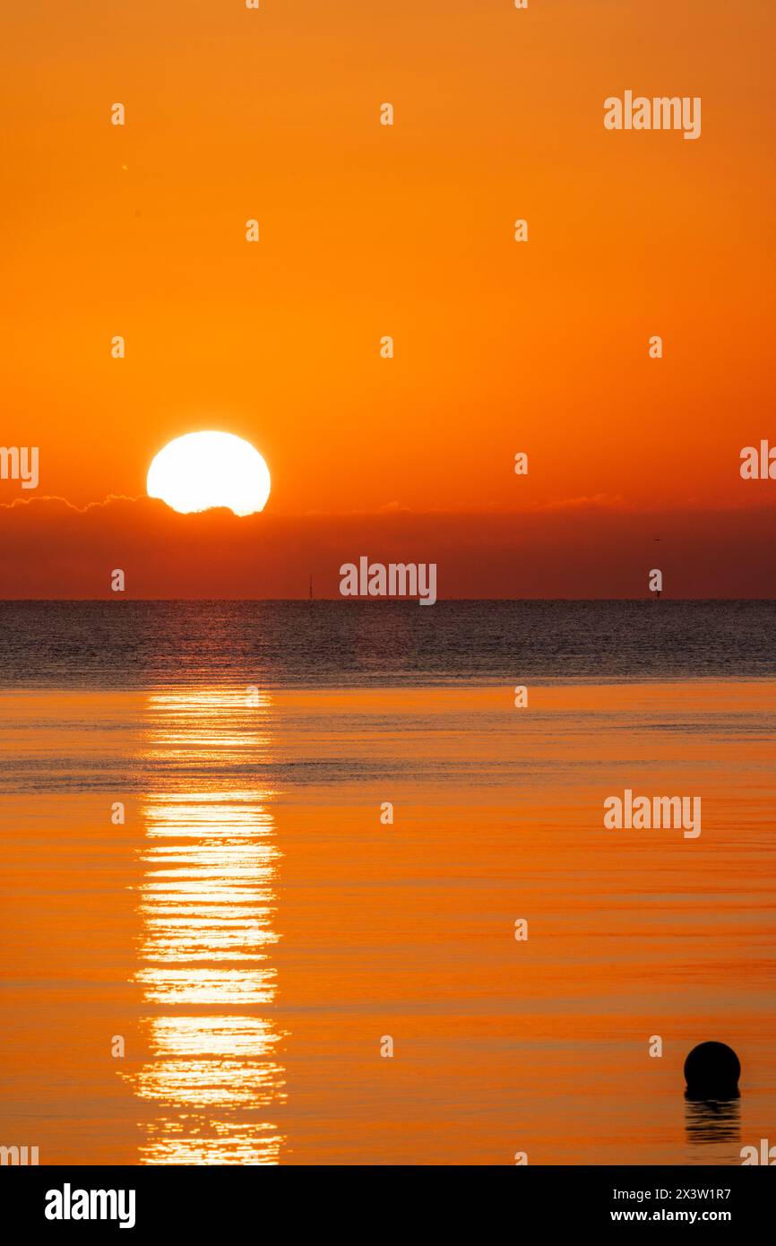 Sonnenaufgang über einem sehr ruhigen Meer in Herne Bay an der Küste von Kent. Dünne Wolkenschicht am Horizont, über der die Sonne scheint, das Meer hat eine orange Farbe. Stockfoto