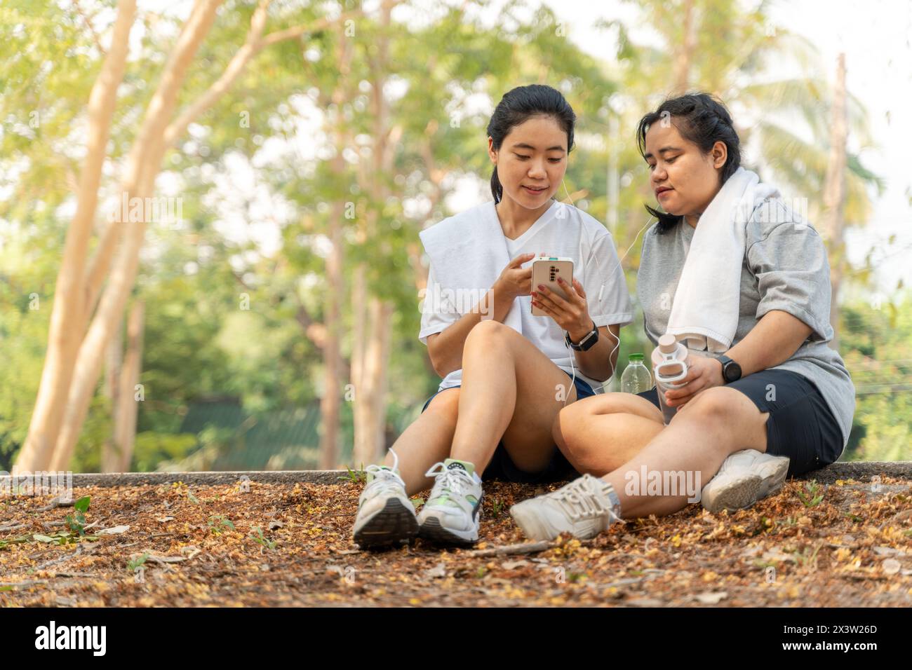 Zwei glückliche asiatische Freundinnen hören Musik von einem Telefon, während sie nach ihrem Morgenlauf zusammen an der kurvenreichen Straße sitzen Stockfoto