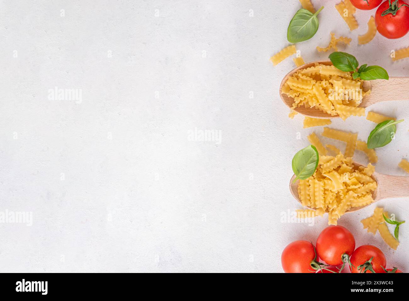 Rohe, ungekochte Mafaldine-Nudeln mit Zutaten zum Kochen. Kurze italienische Mafaldine-Pasta mit Olivenöl, Kirschtomaten, Basilikum und Gewürzen auf weißer Küche Stockfoto