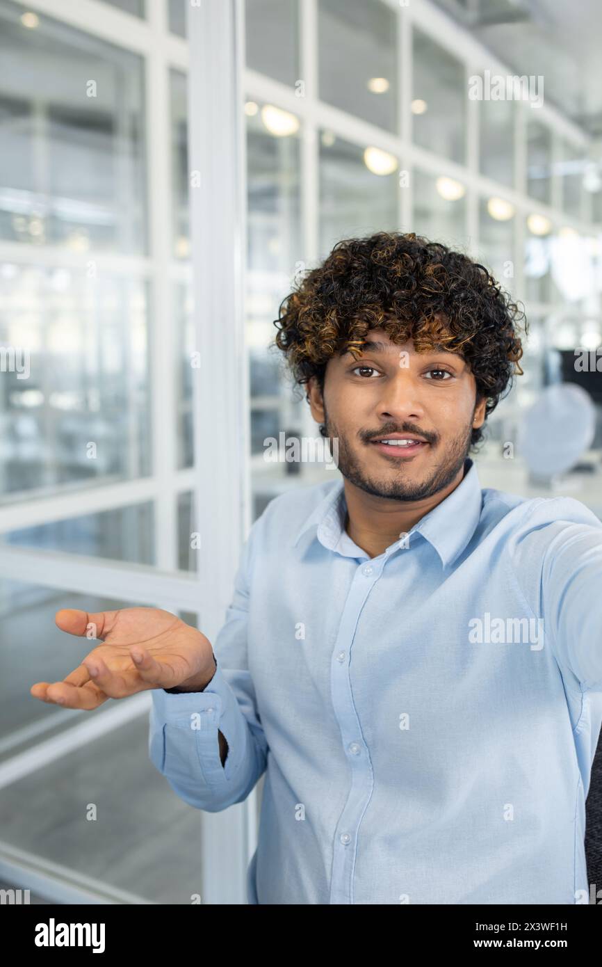 Ein lächelnder junger Mann mit lockigen Haaren macht einen Videoanruf aus einem modernen Büro. Er schaut in die Kamera und führt ein freundliches Gespräch. Stockfoto