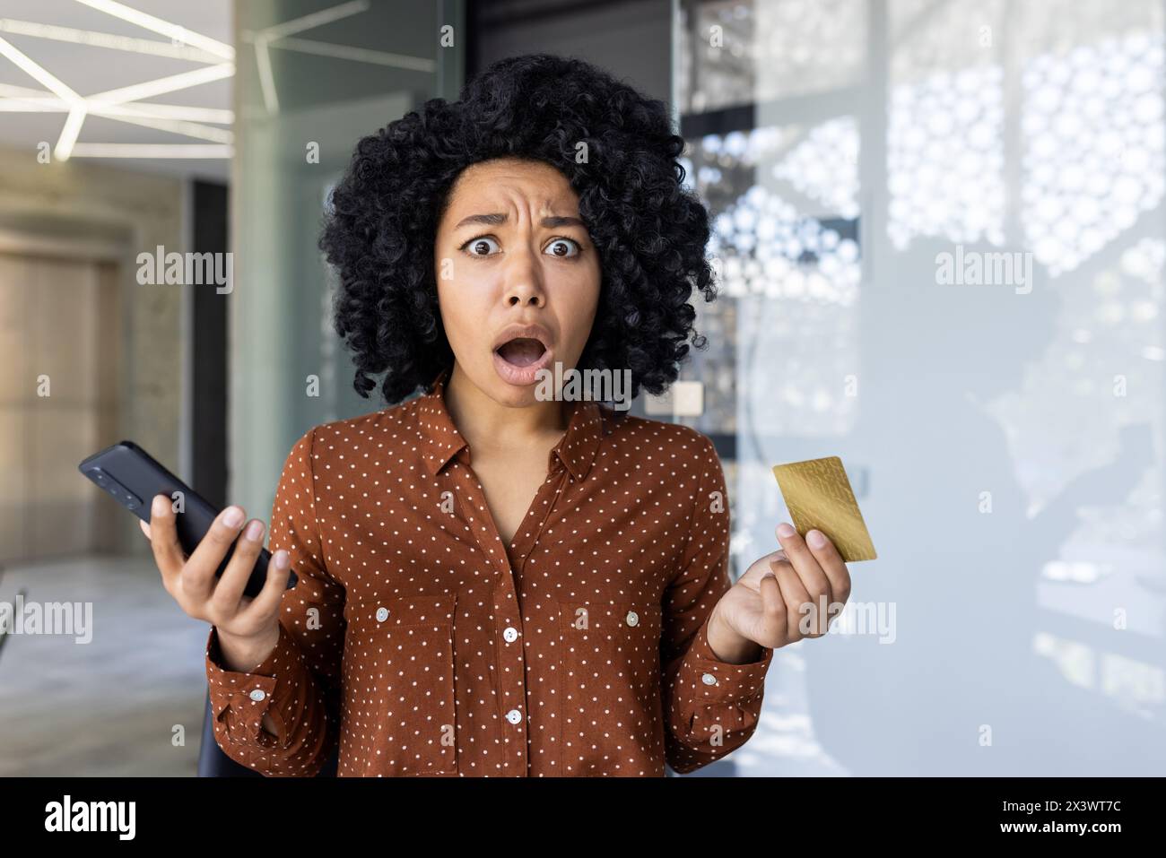 Eine junge Afroamerikanerin scheint schockiert zu sein, während sie ein Smartphone und eine Kreditkarte hält. Ihr Ausdruck deutet auf eine unerwartete Situation hin, die möglicherweise mit Finanzen zusammenhängt. Stockfoto