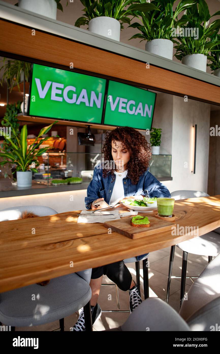 Frau mit lockigen Stühlen in vegetarischem Café oder Restaurant mit grünem Smoothie und Pflanzenmahlzeit auf dem Tisch, das Smartphone sieht aus. Veranstaltungsorte VEGANES Schild Disla Stockfoto