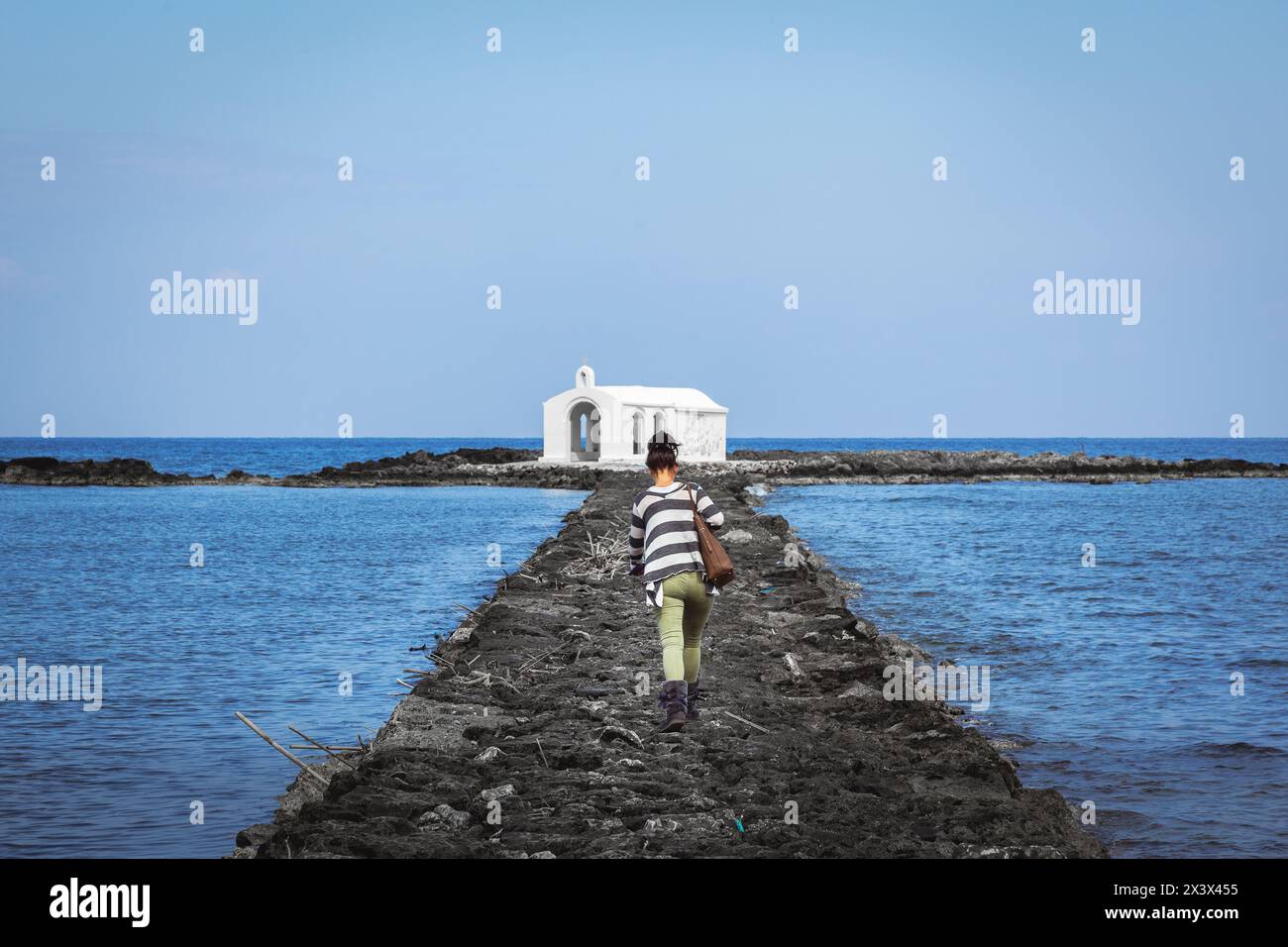 Eine junge kaukasische Frau, die auf dem künstlichen Steinsteg zur auf See gebauten weißen Kapelle des Heiligen Nikolaus (Agios Nikolaos) geht Stockfoto