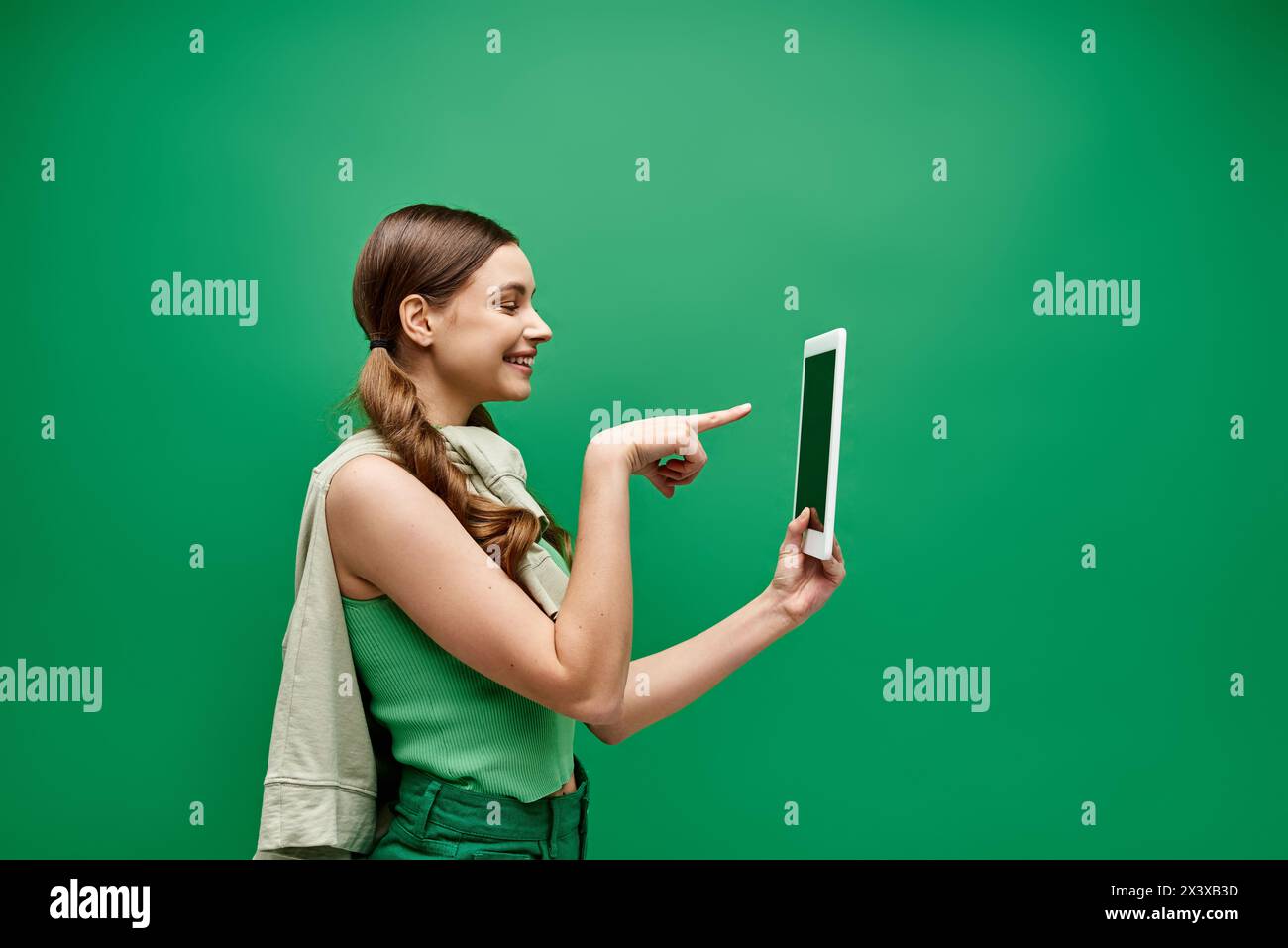 Eine junge Frau im Alter von 20 hält ein Tablet und zeigt auf es in einem Studio mit grünem Hintergrund. Stockfoto