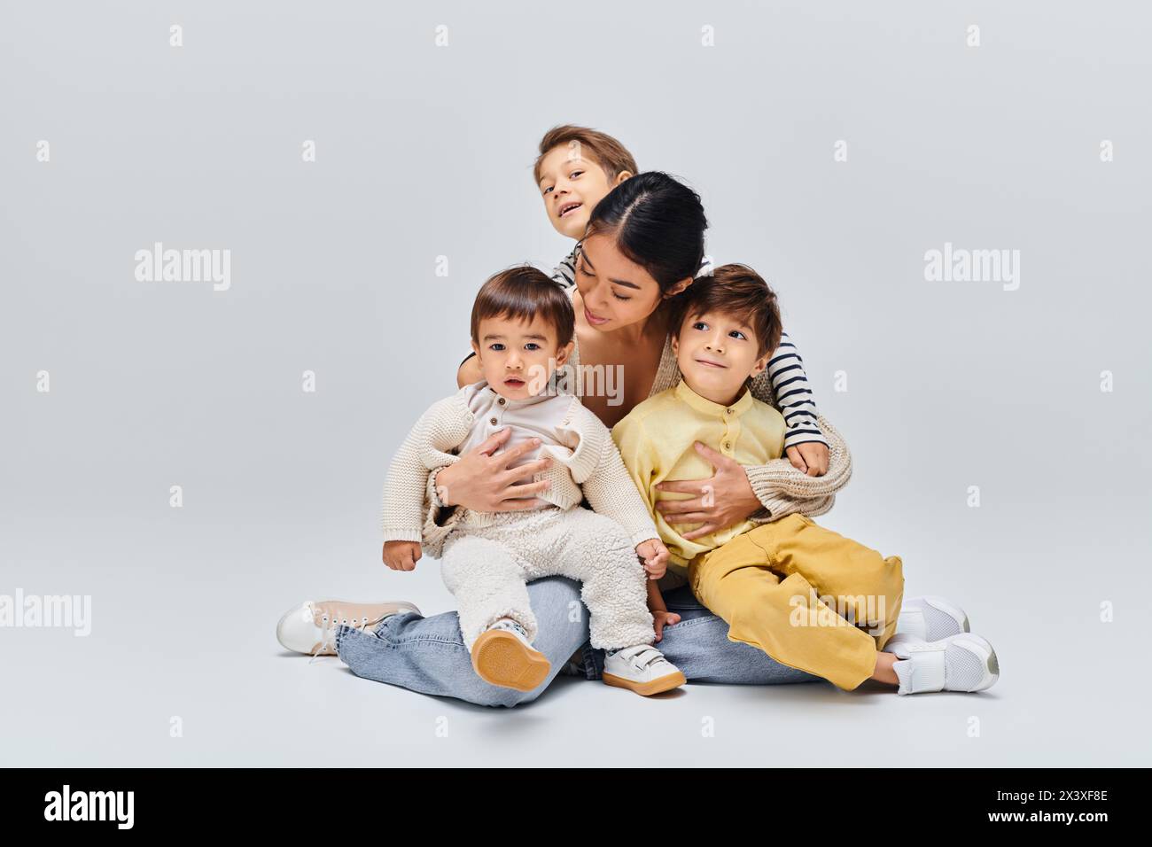 Eine junge asiatische Mutter sitzt mit ihren Kindern auf dem Boden in einem Studio auf grauem Hintergrund. Stockfoto