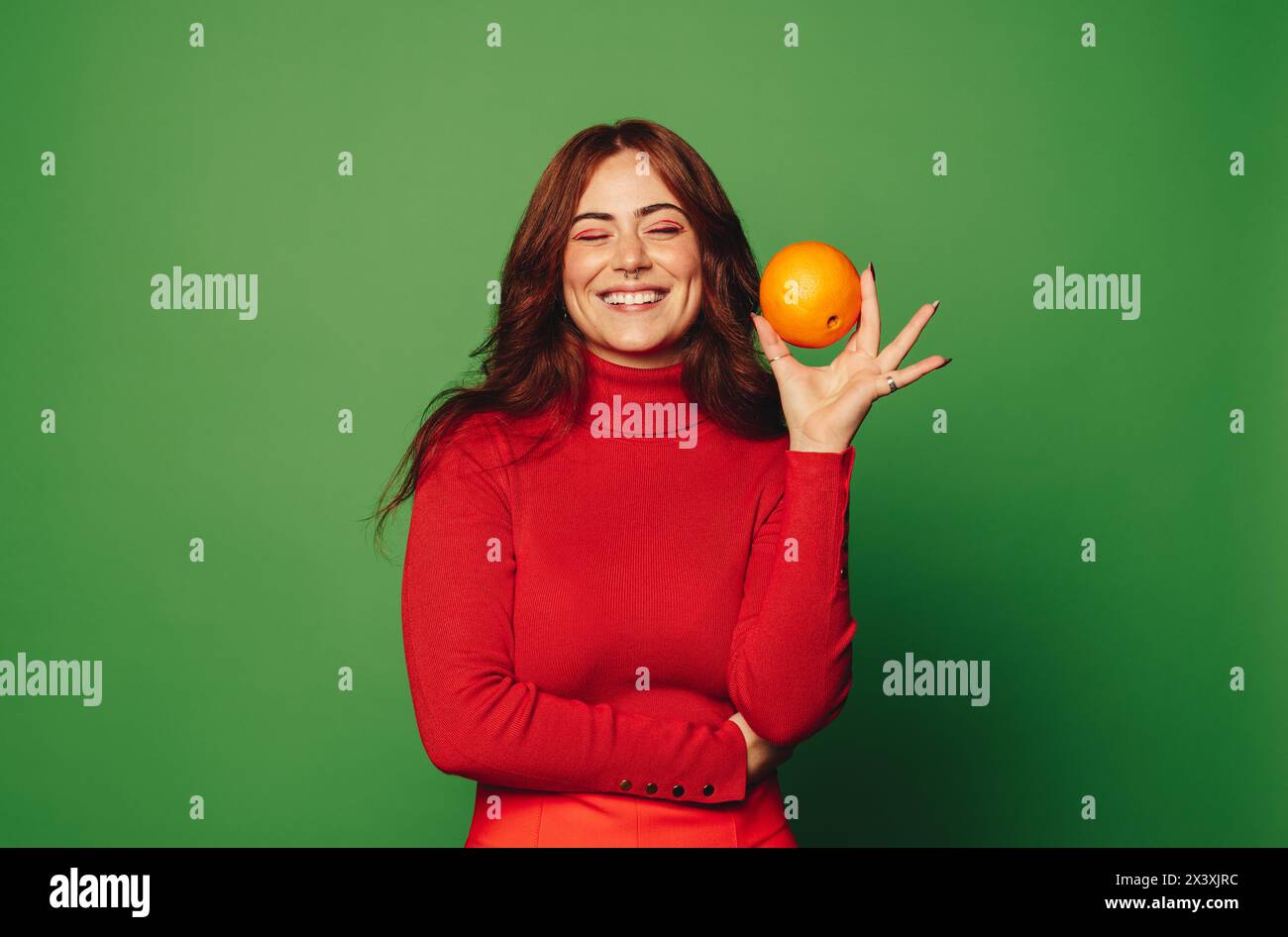 Glückliche Frau in lässiger Kleidung lächelt, während sie in einem Studio mit grünem Hintergrund eine orangene Frucht hält. Die Frucht steht für Frische, Ernährung und Wohlbefinden Stockfoto