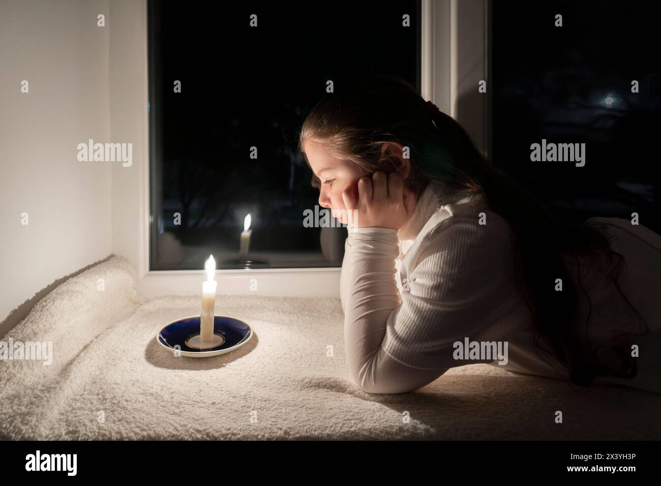 Blackout. Ein Mädchen in einem dunklen Raum liegt auf der Fensterbank neben dem Fenster und schaut sich eine brennende Kerze an. Stromausfall Stockfoto