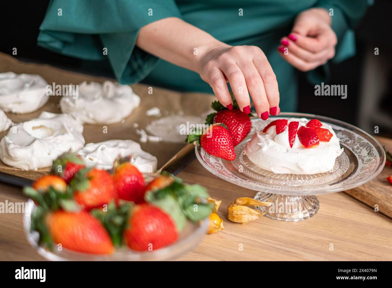 Ein Koch in einer grünen Schürze stellt Erdbeerpawlovas in einer hellen Küche zusammen, mit weiteren Desserts im Hintergrund. Ideal für kulinarische Websites und de Stockfoto