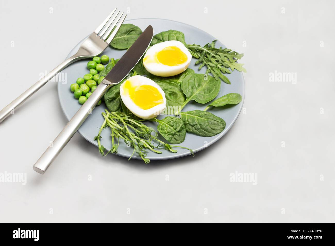 Zwei Hälften gekochtes Hühnerei, Spinatblätter, Rucola und grüne Erbsen auf grauer Keramikplatte. Gabel und Messer auf Platte. Graue Serviette auf dem Tisch. Top-Film Stockfoto