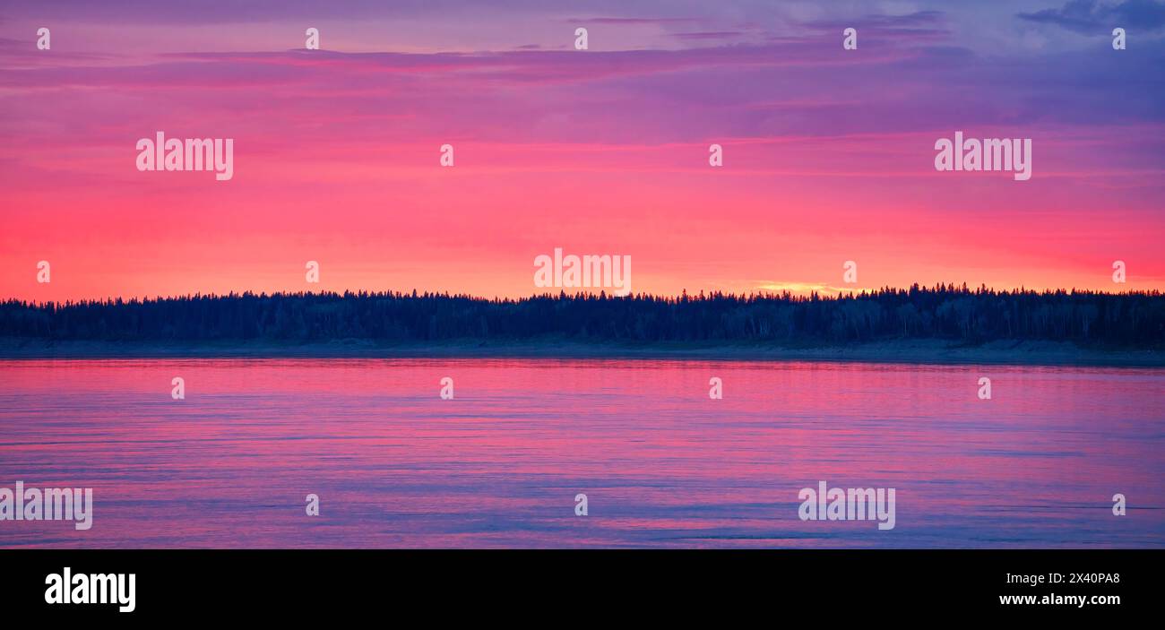 Der Abendhimmel entzündet sich mit Farbe, während die Sonne unter der Horizontlinie in Fort Simpson untergeht. Der Mackenzie River reflektiert die Farbe in einem wunderschönen ... Stockfoto