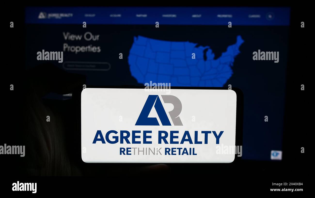 Person, die ein Mobiltelefon mit dem Logo des US-Immobilienunternehmens Agree Realty Corporation vor der Unternehmenswebseite hält. Schwerpunkt auf dem Display des Telefons. Stockfoto