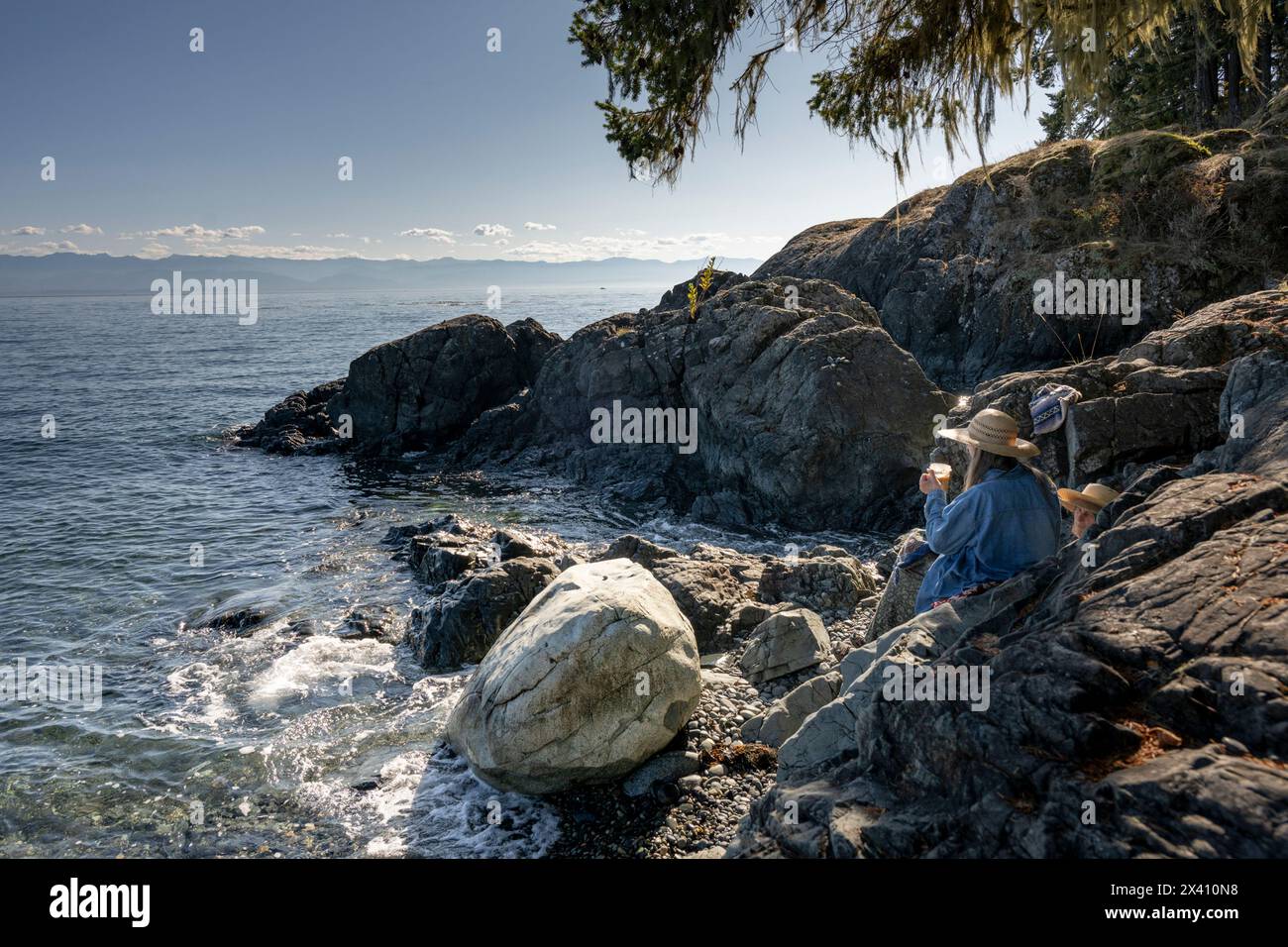 Zwei Personen sitzen an einem Felsenstrand am Wasser und genießen ihren Morgenkaffee, während sie an einem sonnigen Tag auf das Meer blicken, in der Sooke Bay entlang... Stockfoto