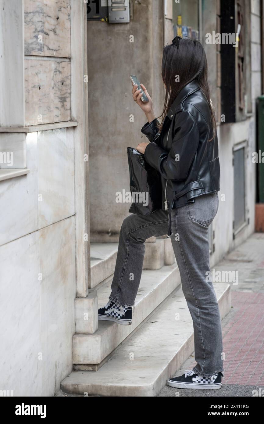 Eine junge Frau hält auf einem Bürgersteig an, um ihr Smartphone zu überprüfen. Sie hält eine Einkaufstasche in der Hand und trägt stilvolle schwarze Kleidung. Athen, Griechenland Stockfoto