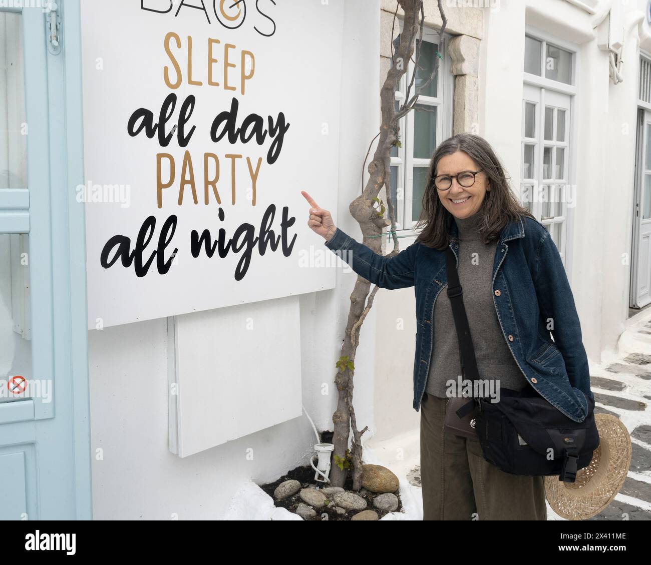 Reife Frau zeigt und lacht über ein jugendliches und sorgloses inspirierendes Schild, auf dem steht "Schlaf den ganzen Tag Party die ganze Nacht", das auf einem weißen Gebäude steht... Stockfoto