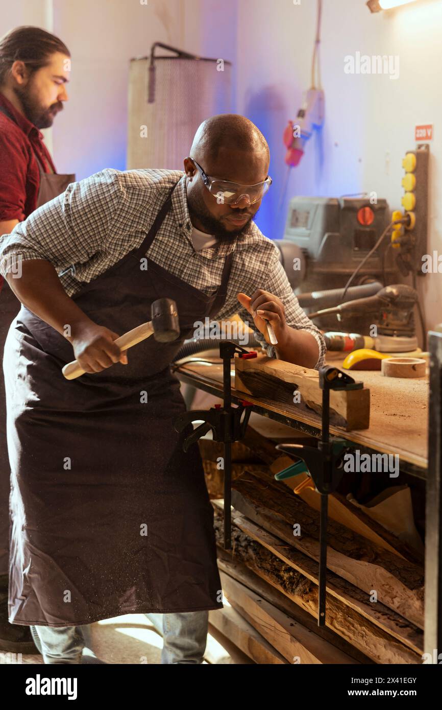 Handwerker schnitzen Entwürfe in Holz mit Meißel und Hammer neben dem Kollegen. Handwerkliches Formen von Holzteilen mit Schutzausrüstung zur Vermeidung von Arbeitsunfällen, unterstützt durch den Auszubildenden Stockfoto
