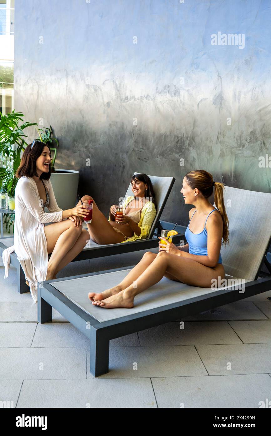 Drei Frauen sitzen auf Liegestühlen und trinken Saft. Sie lächeln und genießen die Gesellschaft des anderen. Vertikale Aufnahme Stockfoto