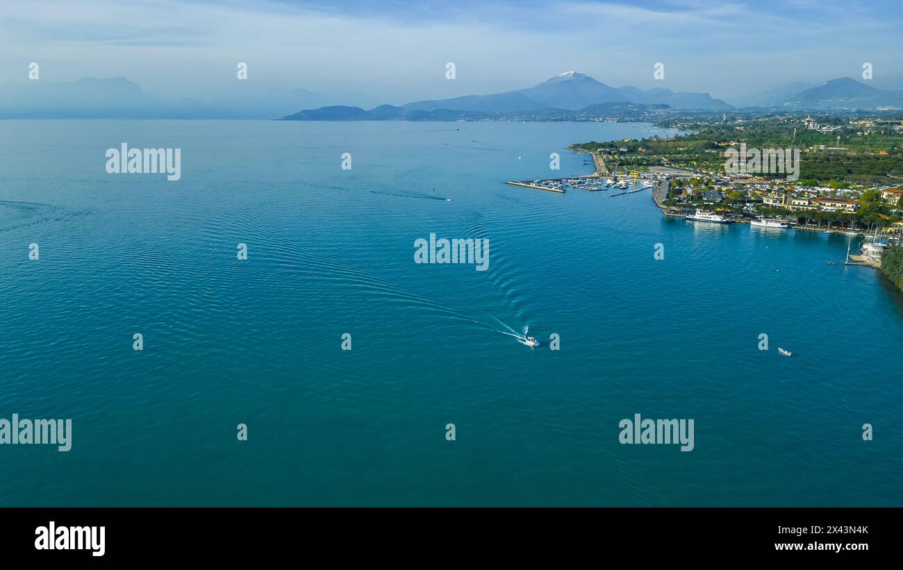 Mit Blick auf das ruhige Wasser des Gardasees, mit Booten, die an der malerischen Stadt Peschiera del Garda vor bergiger Kulisse vorbeisegeln. Hohe Qualität Stockfoto