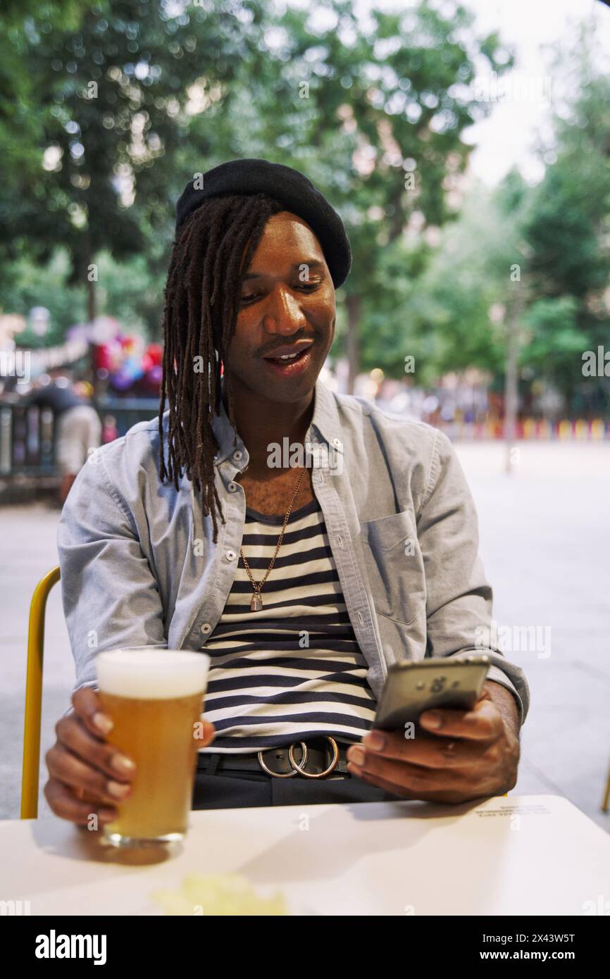 Ein lächelnder Mann sitzt im Restaurant, trägt ein schickes, lässiges Outfit, benutzt ein Telefon und trinkt ein Bier. Stockfoto