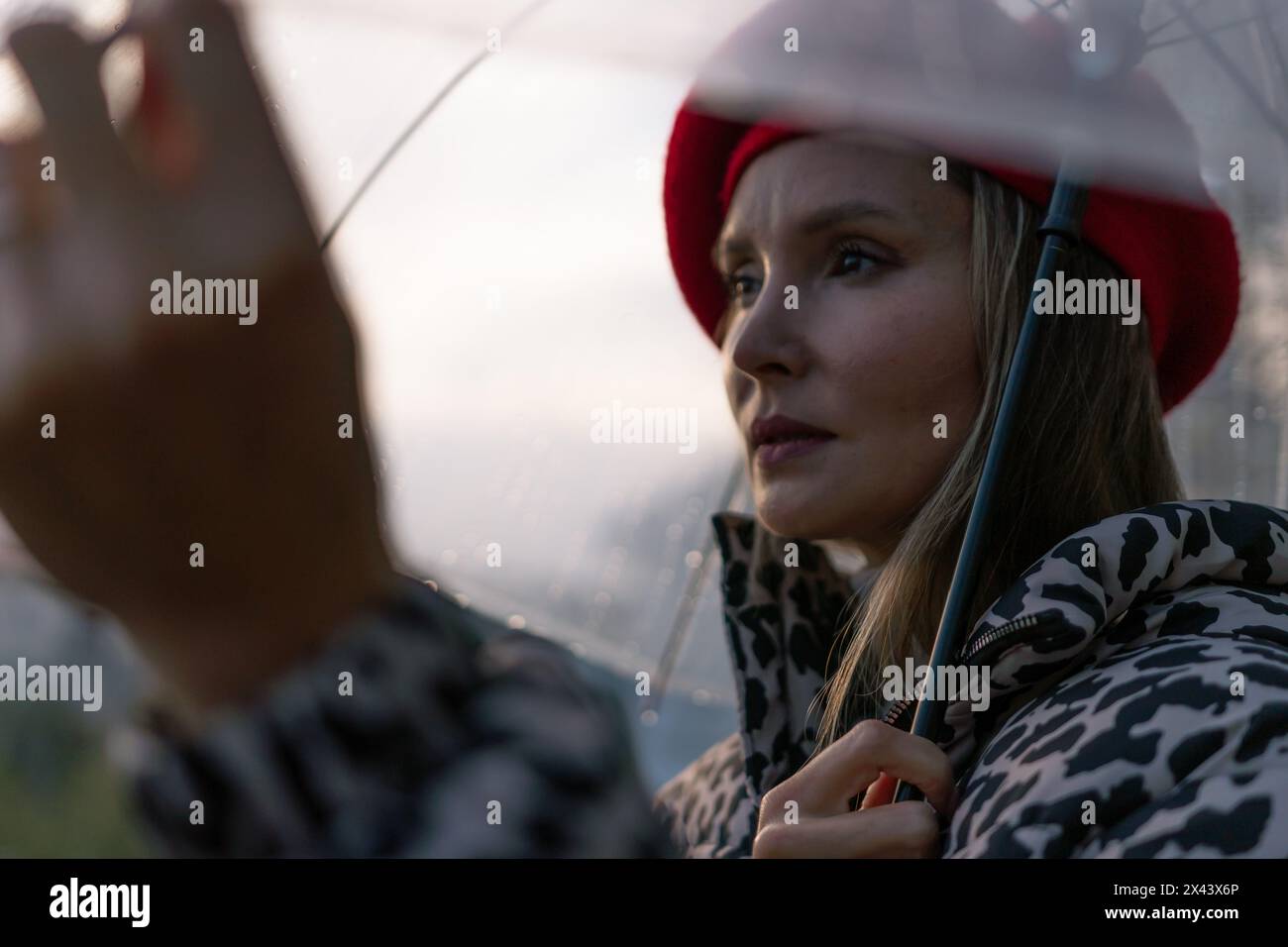 Eine Frau, die einen roten Hut und einen Mantel mit Leopardenmuster trägt, hält einen klaren Regenschirm. Der Schirm ist offen und die Frau blickt in den Himmel. Die Stockfoto