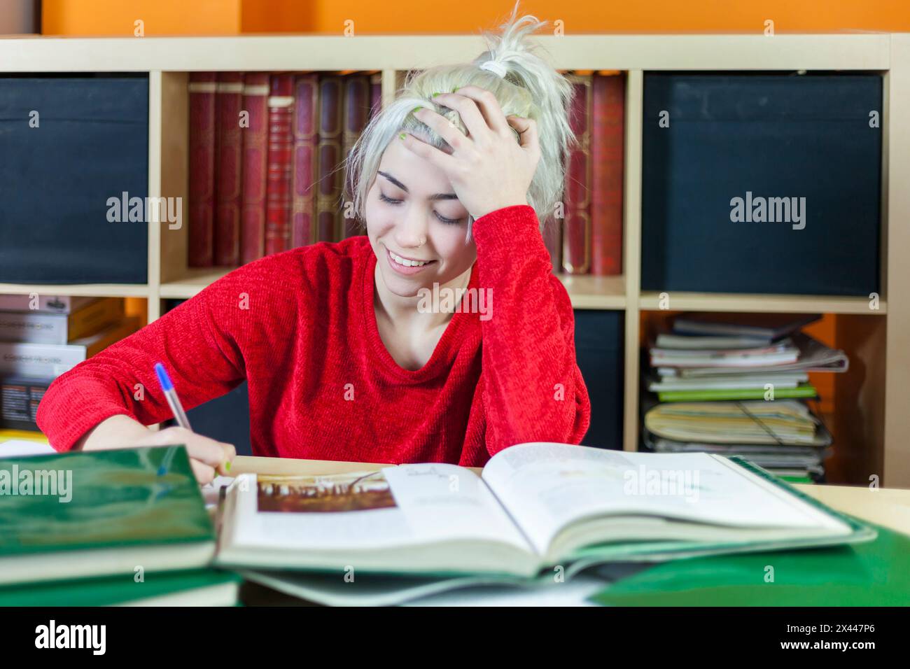 Junge Frau mit blonden Haaren, lächelt beim Schreiben von Notizen. Einen roten Pullover tragen, mit Büchern an der Bücherei lernen, eine fröhliche Stimmung vermitteln Stockfoto