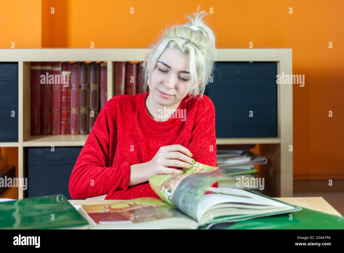 Junge Frau mit gebleichtem blondem Haar, mit Fokus auf Lesen. Rot gekleidet, an einem Bücherpult sitzend, umgeben von Büchern, symbolisiert tiefes Konzentra Stockfoto