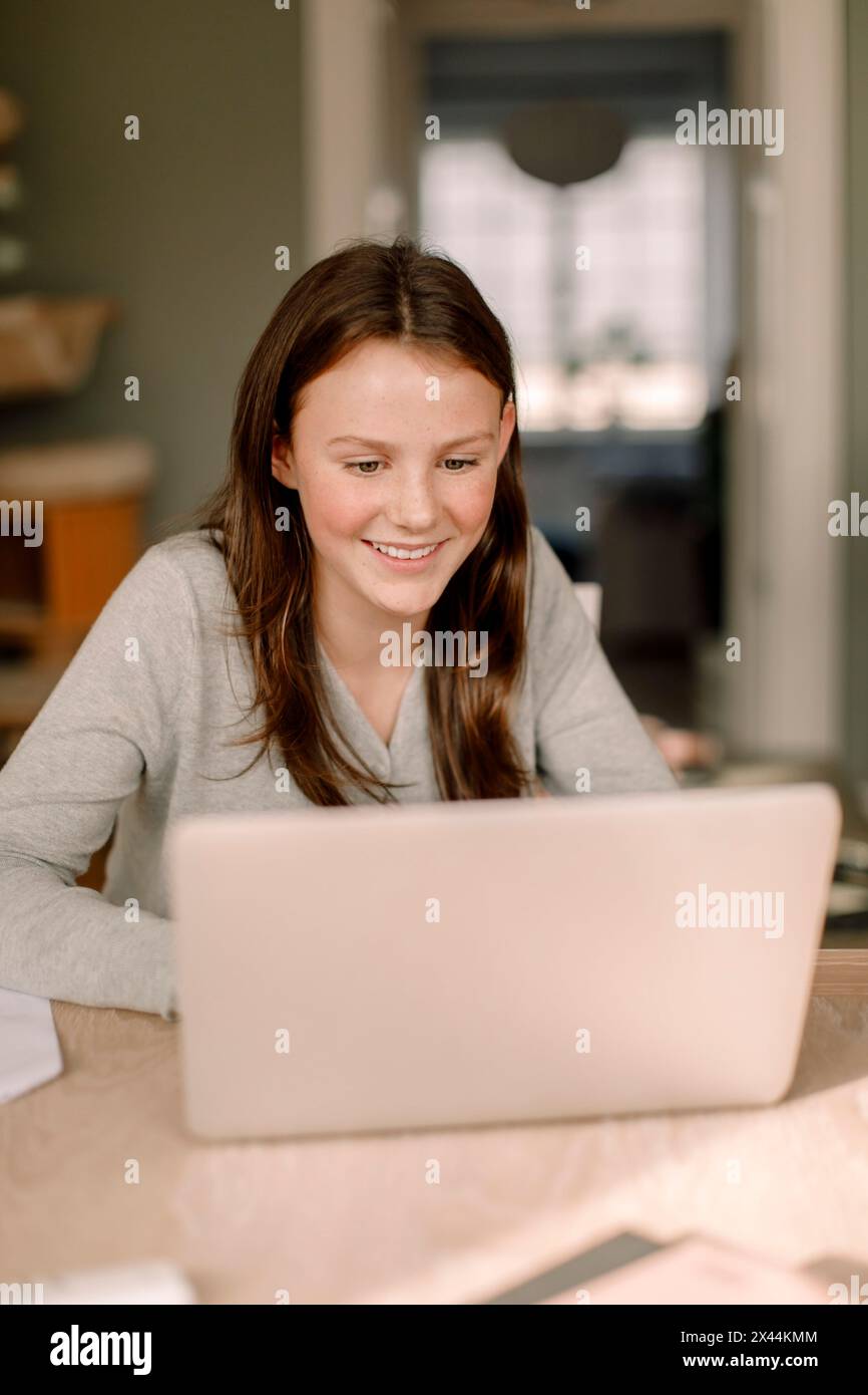 Lächelndes Teenager-Mädchen, während er den Laptop am Esstisch in der Küche benutzte Stockfoto