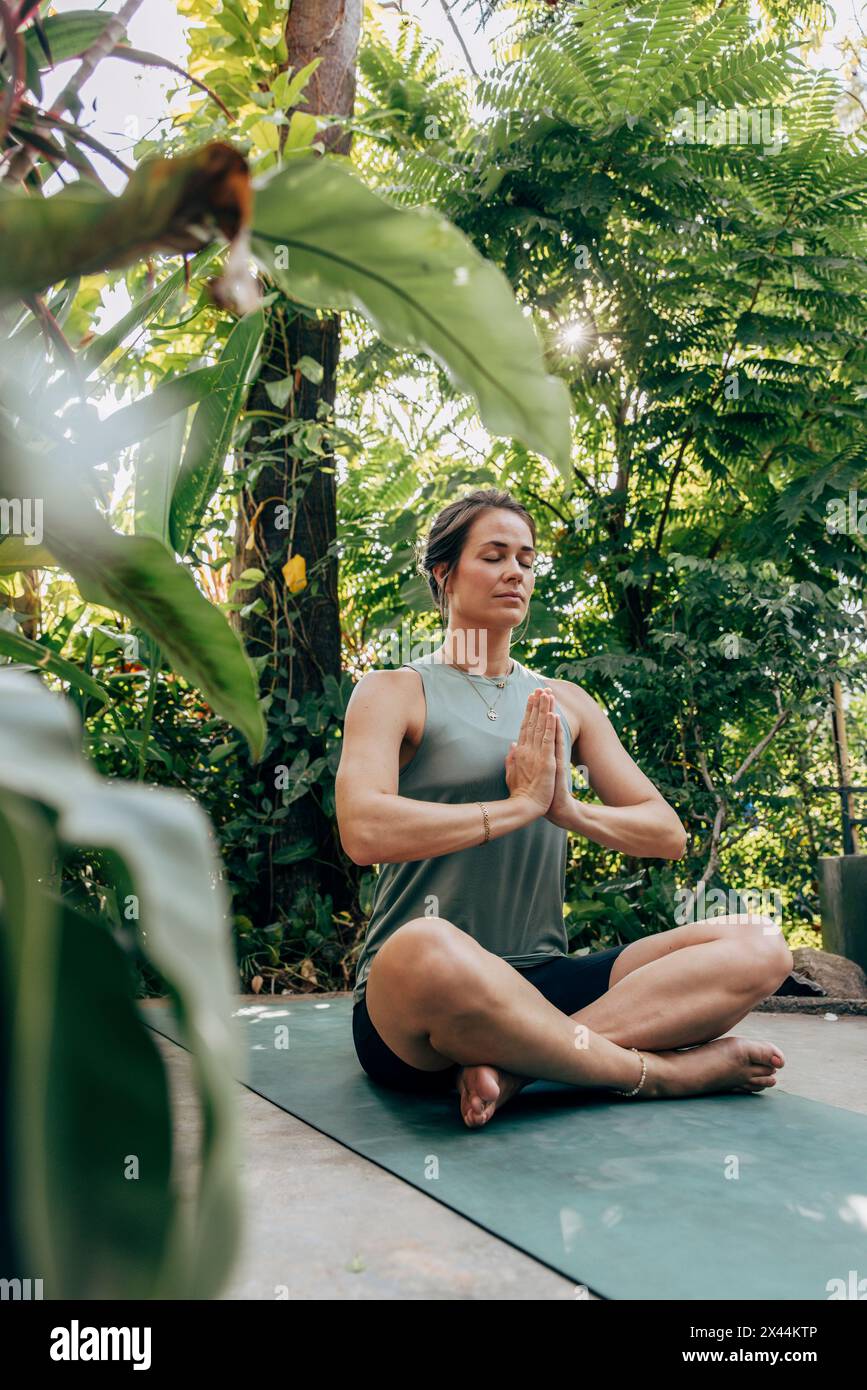 Reife Frau übt Meditation, während sie im Kreuzbein inmitten von Pflanzen im Wellness-Resort sitzt Stockfoto