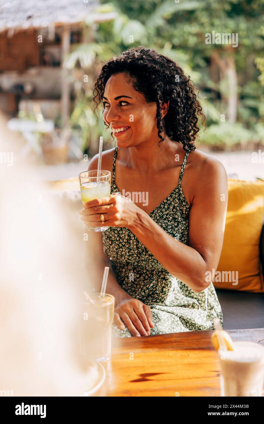 Lächelnde Frau mit lockigem Haar, die frischen Saft genießt, während sie im Wellnessresort sitzt Stockfoto