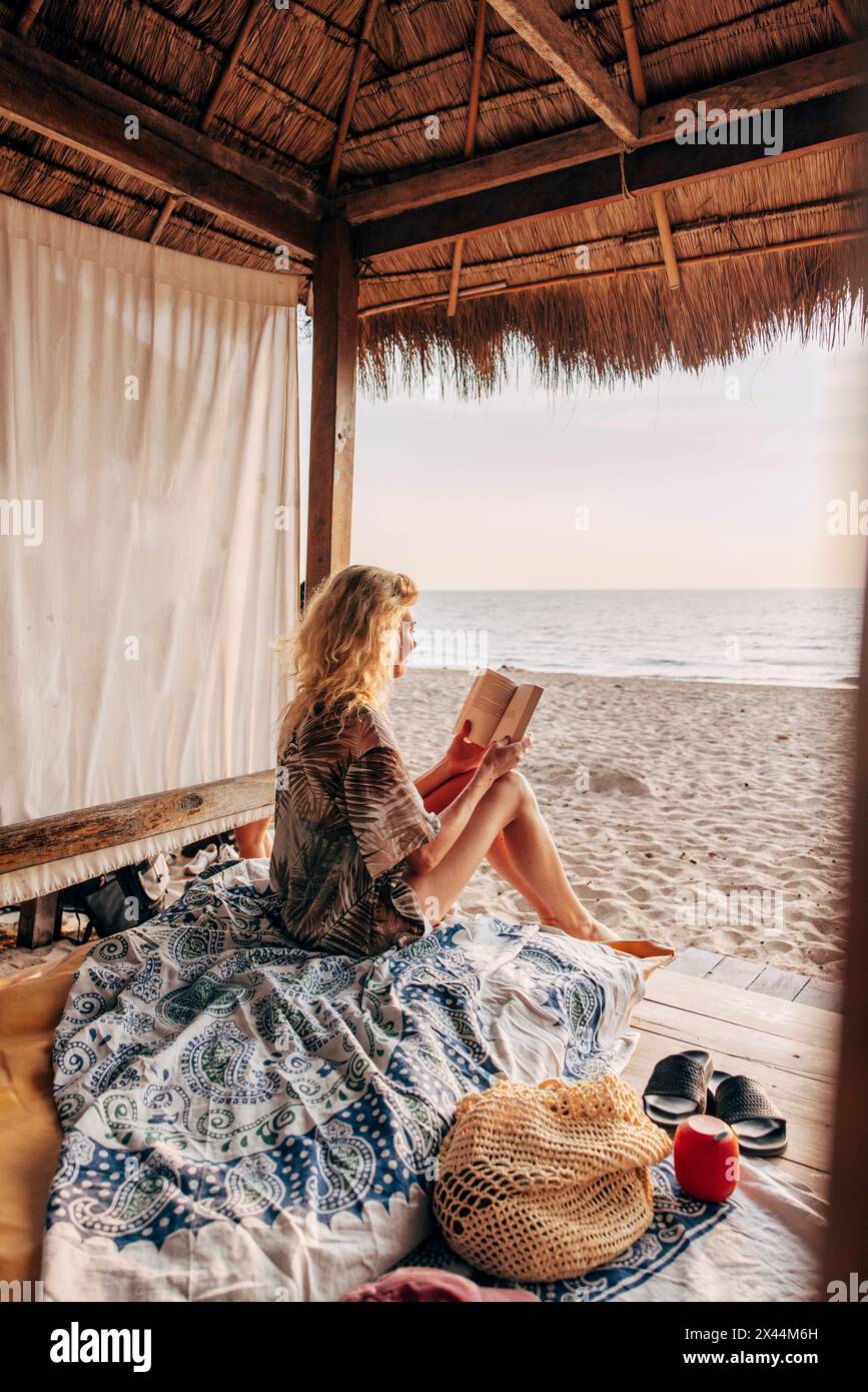 Frau liest Buch, während sie im Urlaub am Strand in der Cabana sitzt Stockfoto