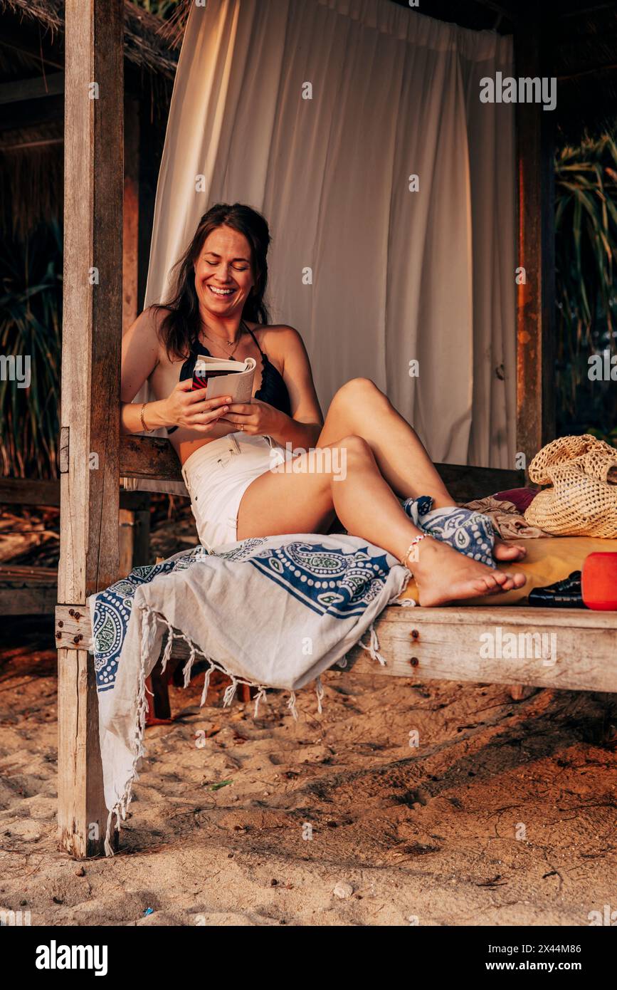 Glückliche Frau, die ein Buch liest, während sie im Urlaub am Strand in der Cabana sitzt Stockfoto
