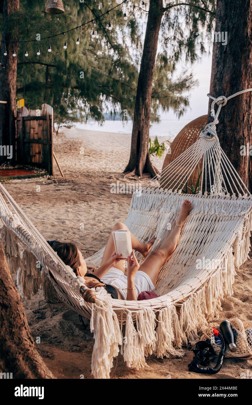 Frau liest Buch, während sie sich in der Hängematte im Urlaub am Strand liegend Stockfoto