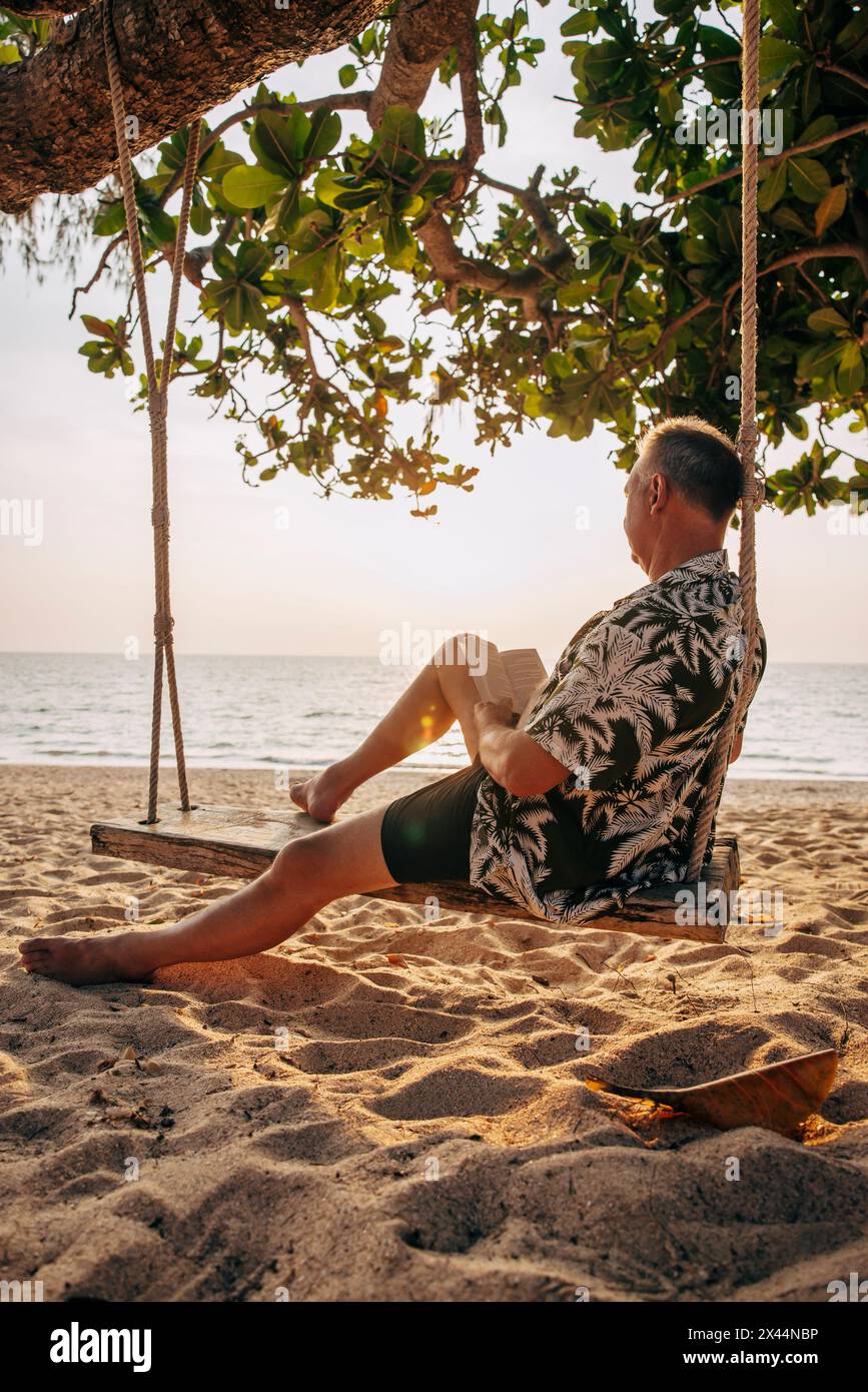 Seitenansicht eines älteren Mannes, der ein Buch liest, während er am Strand auf einer Schaukel saß Stockfoto
