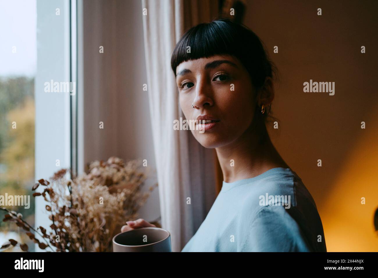 Seitenporträt einer jungen Frau, die zu Hause eine Kaffeetasse in der Nähe eines Fensters hält Stockfoto