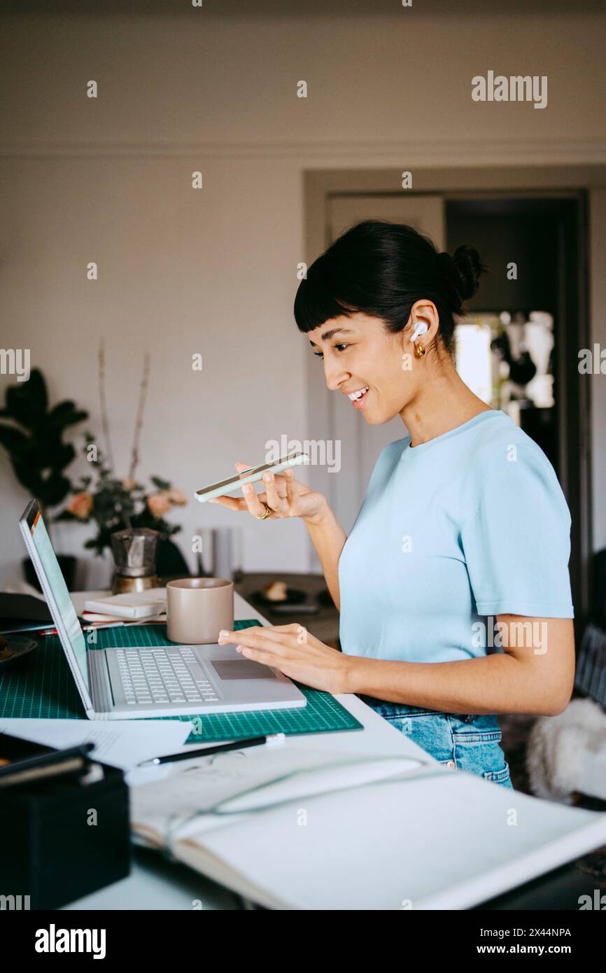 Lächelnde junge Freiberuflerin, die über Lautsprechertelefon spricht, während sie im Heimbüro einen Laptop benutzt Stockfoto