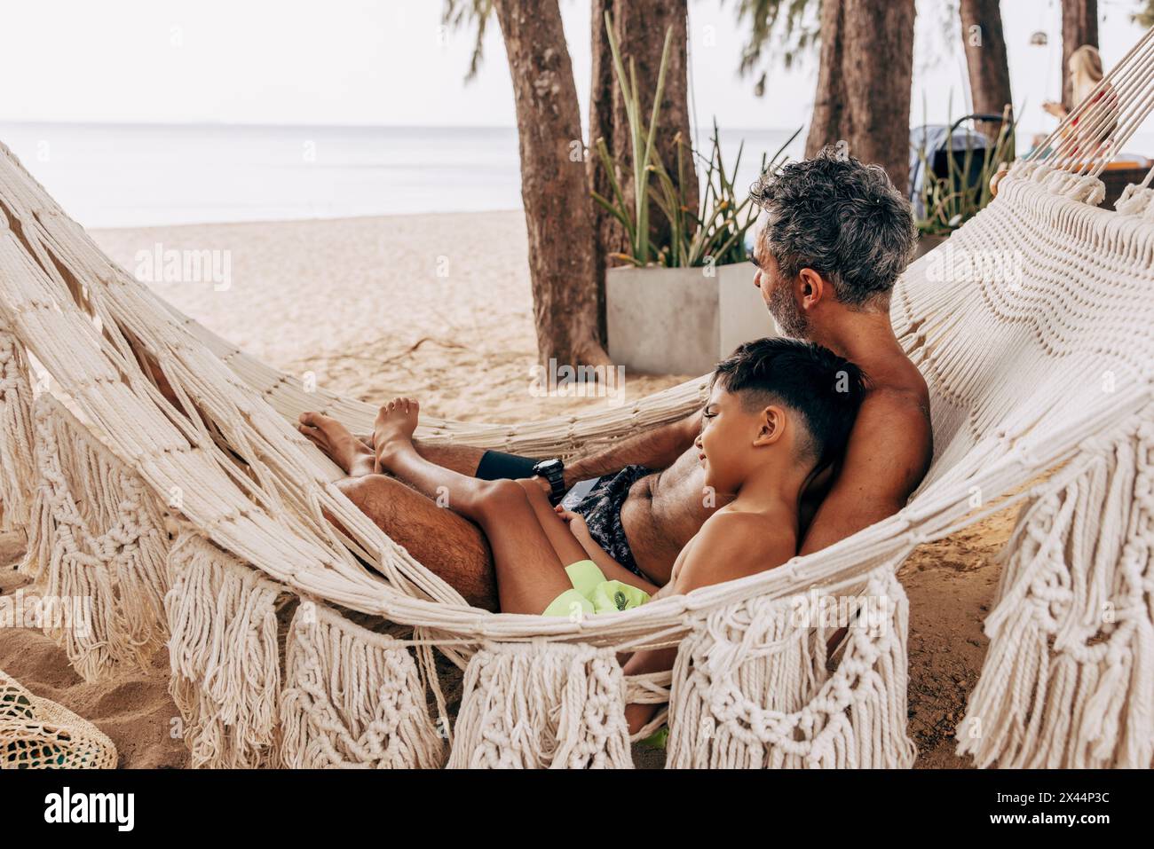 Vater und Sohn ohne Hemd, die sich im Urlaub in der Hängematte am Strand entspannen Stockfoto